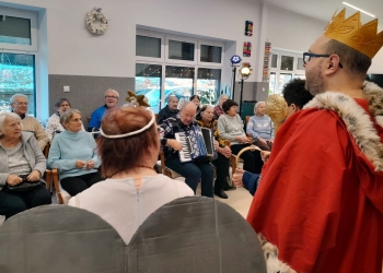 Kolędnicy ubrani w strój anioła i czerwony płaszcz z koroną na głowie. W tle seniorzy siedzący na krzesłach. Jeden z seniorów gra na akordeonie.