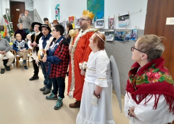 Na pierwszym planie uczestnicy kolędowania ubrani w stroje kolędnicze.Kobieta ma na ramionach kolorową chustę ludową, obok osoba ubrana jest w biały strój i skrzydła anioła, meżczyzna w czerwonym płaszczu ma na głowie koronę, na dalszym planie seniorzy. 