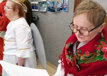 Uczestniczka kolędowania trzyma w rękach nuty, na ramionach ma kolorową chuste ludową. Na dalszym planie jest osoba w stroju anioła.