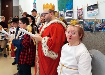 Na pierwszym planie uczestnicy kolędowania ubrani w stroje kolędnicze.Kobieta ma na sobie biały strój i skrzydła anioła, meżczyzna w czerwonym płaszczu ma na głowie koronę, na dalszym planie seniorzy. 