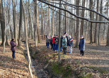 Grupa osób stoi wzdłuż strumyka, jedna osoba stoi po przeciwnej stronie rowu, przodem do grupy, coś opowiada. Osoby znajdują się w lesie.