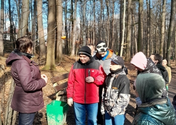 Osoby na zdjęciu uczestniczą w wycieczce ekologicznej w lesie. mają na sobie ciepłą odzież.