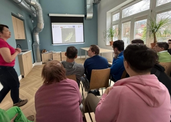 Zdjęcie przedstawia osoby biorące udział w zajęciach edukacyjnych. Uczestnicy siedza w rzędach na krzesłach, skupiając swoją uwagę na osobie prowadzącej, która stoi po lewej stronie.