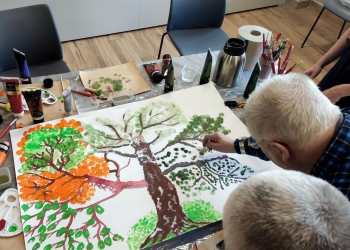Osoby skupione na wspólnej pracy. Tworzą rysunek na kartonie leżącym na stole.