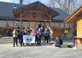 Na zdjęciu widać grupę kilkunastu osób stojących przed schroniskiem. Grupa jest ubrana w odzież turystyczną. Osoby trzymają flagę z logo klubu górskiego.