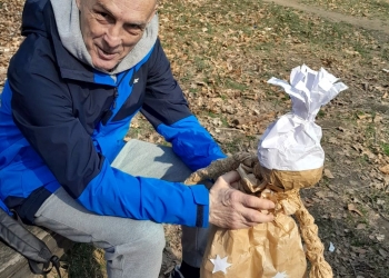 Na zdjęciu widać mężczyznę siedzącego na drewnianej ławce w lesie. Ubrany jest w sportową kurtkę w odcieniach niebieskiego i szarości, dresowe spodnie oraz sportowe buty. Patrzy w stronę aparatu i lekko się uśmiecha. Obok niego znajduje się wykonana z papieru Marzanna.