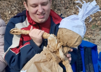Na zdjęciu widać osobę siedzącą w lesie i trzymającą w dłoniach papierowa kukłę Marzanny. 