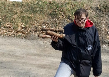 Na zdjęciu widać młodego mężczyznę pozującego do fotografii na tle budynku schroniska. Meżczyzna trzyma w dłoniach przygotowane drewno do ogniska.