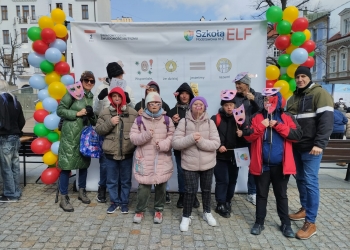 Na zdjęciu znajdują się osoby świętujące Światowy Dzień Świadomości Autyzmu. W tle jest baner nawiązujący do tego wydarzenia oraz kolorowe balony.