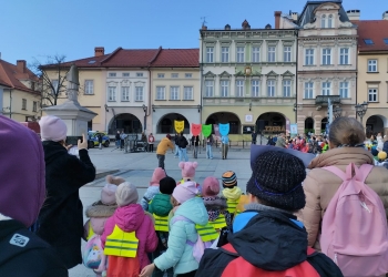 Zdjęcie przedstawia osoby zgromadzone na Rynku podczas wydarzenia. Na dalszym planie kilka osób tzryma maski symbolizujące różne emocje. W tle kamienice.