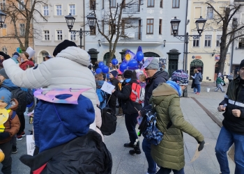 Zdjęcie przedstawia osoby biorące udział w marszu. W rękach trzymają niebieskie baloniki. W tle kamienice.
