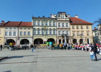 Zdjęcie przedstawia osoby zgromadzone na Rynku podczas wydarzenia. Na dalszym planie kilka osób tzryma maski symbolizujące różne emocje. W tle kamienice.