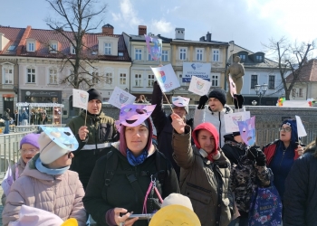 Na zdjęciu znajdują się osoby świętujące Światowy Dzień Świadomości Autyzmu. W tle jest baner nawiązujący do tego wydarzenia oraz kolorowe balony.W tle kamienice.