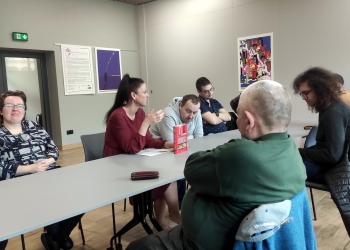 Grupa osób siedzi dookoła szarego stołu, jedna z osób prowadzi wykład, reszta grupy słucha. 