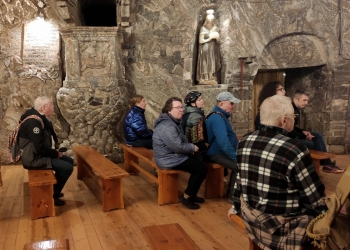 Uczestnicy wycieczki znajdują się w kaplicy, siedzą na niskich ławkach. W tle ściana, na której znajdują się solne rzeźby świętych.