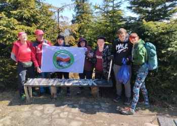 Na zdjęciu widać grupę siedmiu osób stojących na leśnej polanie wśród gęstych, zielonych drzew iglastych. Większość ma sportowe ubrania, czapki i plecaki. Część grupy trzyma biały baner z kolorowym logo. Przed nimi znajduje się drewniana ławka z napisem „Klimczok”. Osoby stoją blisko siebie, niektóre patrzą w obiektyw, inne lekko w bok. Po prawej stronie widać kobietę w okularach przeciwsłonecznych i zielonej kurtce, która uśmiecha się i ma na plecach duży plecak. Jedna z osób trzyma worek, prawdopodobnie ze śmieciami lub sprzętem.. .