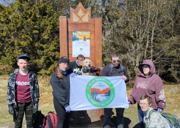 Na zdjęciu widać grupę osób stojących na polanie wśród gęstych, zielonych drzew iglastych. Większość ma sportowe ubrania, czapki i plecaki. Część grupy trzyma biały baner z kolorowym logo. Przed nimi znajduje się drewniana ławka z napisem „Klimczok”. Osoby stoją blisko siebie, niektóre patrzą w obiektyw, inne lekko w bok.  Na ziemi leżą worki, prawdopodobnie ze śmieciami.