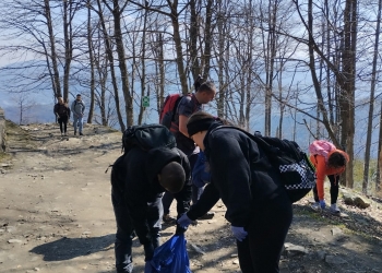 Zdjęcie przedstawia grupę osób zaangażowanych w sprzątanie szlaku górskiego. Na pierwszym planie dwie osoby pochylają się nad dużym workiem na śmieci, jedna z osób ma rękawiczki ochronne.  W tle widać więcej drzew i otwartą przestrzeń.