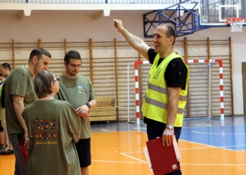 Na zdjęciu widać grupę osób znajdujących się w hali sportowej. Po prawej stronie stoi mężczyzna w żółtej kamizelce odblaskowej,uśmiecha się i wskazuje ręką w górę, jakby udzielał instrukcji albo kierował uczestnikami. W drugiej ręce trzyma czerwoną teczkę i dokumenty. Przed nim stoją trzy młodsze osoby ubrane w zielone koszulki sportowe. Jedna z nich jest odwrócona tyłem; na koszulce widoczny jest napis „ŚDS Podkowa w Bielsku-Białej”. Pozostali słuchają prowadzącego z uwagą. W tle widać elementy wyposażenia hali: kosz do koszykówki, bramkę sportową oraz drewniane drabinki gimnastyczne. 