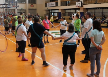 Na zdjęciu widać grupę osób stojących w kręgu na hali sportowej. Uczestnicy trzymają się za ręce. Większość osób ubrana jest w stroje sportowe, a przez ramię mają przewieszone niebieskie szarfy. W tle widać więcej uczestników wydarzenia, organizatorów w żółtych kamizelkach odblaskowych oraz osobę przebraną za dużą maskotkę psa Reksia.  Hala jest dobrze oświetlona i wyposażona w kosze do koszykówki oraz trybuny, na których siedzą obserwatorzy.
