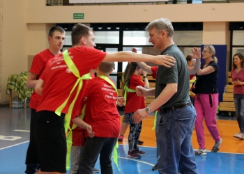 Na zdjęciu widać kilka osób ubranych w czerwone koszulki i jaskrawe zielone szarfy wykonuje ćwiczenie lub zabawę ruchową, stojąc blisko siebie i wyciągając ręce.W centrum kadru znajduje się mężczyzna w ciemnej koszulce i dżinsach, który uśmiecha się i bierze udział w aktywności razem z grupą młodych uczestników. W tle można zauważyć kolejne osoby ćwiczące lub obserwujące wydarzenie.