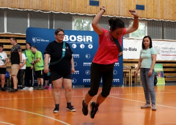 Na zdjęciu uchwycono uczestniczkę zawodów sportowo-integracyjnych w momencie skoku na hali gimnastycznej. Kobieta ubrana w różową koszulkę, czarne legginsy i sportowe buty unosi się nad podłogą z rękami podniesionymi do góry. Przez ramię ma przewieszoną zieloną szarfę z numerem startowym.  W tle stoją inni uczestnicy oraz organizatorzy wydarzenia. Widoczne są również banery z logo „BBOSiR”, napisem „Miasto Bielsko-Biała” oraz „Ministerstwo Sportu i Turystyki”.