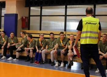 Na zdjęciu widać grupę uczestników siedzących na drewnianej ławce. Większość osób ma na sobie oliwkowozielone koszulki oraz pomarańczowe szarfy z numerami startowymi. Przed grupą stoi organizator w żółtej kamizelce odblaskowej z napisem „Środowiskowy Dom Samopomocy ‘Podkowa’”.
