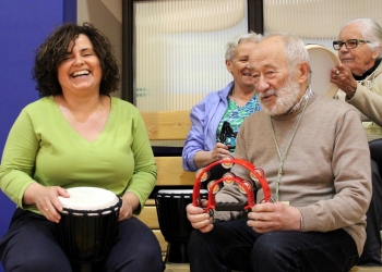 Na zdjęciu widać grupę osób siedzących w hali sportowej i wspólnie grających na instrumentach perkusyjnych. Na pierwszym planie uśmiechnięta kobieta w zielonej bluzce trzyma bęben djembe, a obok niej starszy mężczyzna gra na czerwonym tamburynie. W tle siedzą dwie starsze kobiety – jedna również trzyma instrument. Atmosfera jest radosna i pełna energii.