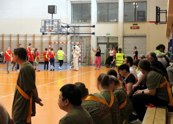 Na zdjęciu widać halę sportową. Na pierwszym planie uczestnicy w oliwkowych koszulkach z pomarańczowymi szarfami siedzą na ławkach i obserwują przebieg zawodów. Część osób rozmawia lub przygląda się temu, co dzieje się na boisku.  W tle grupa uczestników w czerwonych koszulkach przechodzi przez halę, a obok nich stoi osoba w stroju psa Reksia. Widoczni są także organizatorzy w żółtych kamizelkach odblaskowych oraz osoby robiące zdjęcia lub kibicujące.