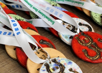 Na zdjęciu widać kilka ceramicznych medali ułożonych na drewnianej powierzchni. Medale mają okrągły kształt i są wykonane w różnych kolorach.Na medalach znajduje się motyw podkowy oraz litery „BB”. Do medali przymocowane są białe wstążki z nadrukiem w kolorze zielonym i niebieskim. Na tasiemkach można zauważyć napis „Środowiskowy Dom Samopomocy Podkowa oraz rok 2026.