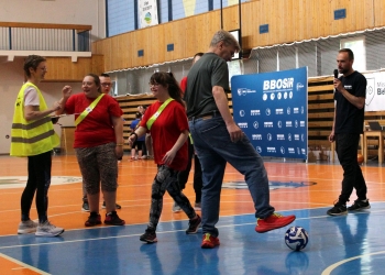 W centrum mężczyzna w jeansach i ciemnej koszulce prowadzi piłkę, stawiając stopę na futbolówce. Obok niego znajdują się uczestniczki w czerwonych koszulkach z zielonymi szarfami, które z uśmiechem obserwują przebieg zadania.  Po lewej stronie stoi organizatorka w żółtej kamizelce odblaskowej, trzymająca kartkę lub notatnik. Po prawej stronie widoczny jest prowadzący wydarzenie z mikrofonem. W tle znajdują się banery BBOSiR oraz elementy hali sportowej.
