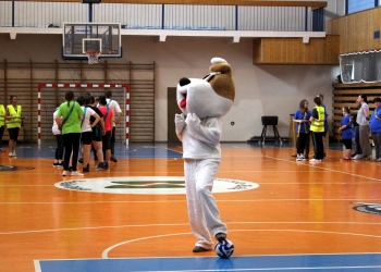 Na zdjęciu widać halę sportową. W centrum boiska stoi osoba przebrana za dużą maskotkę psa reksia, która pozuje z piłką nożną pod stopą. Maskotka wygląda zabawnie i przyciąga uwagę uczestników. W tle znajdują się grupy osób ustawionych w drużynach, część z nich rozmawia lub czeka na swoją kolej do konkurencji. Widoczni są także organizatorzy w żółtych kamizelkach odblaskowych. Nad boiskiem znajdują się kosze do koszykówki oraz bramka do piłki ręcznej.