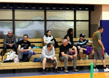 Na zdjęciu widać grupę osób siedzących na drewnianych ławkach w hali sportowej. Część z nich obserwuje wydarzenia odbywające się na boisku, a inni rozmawiają ze sobą. Na pierwszym planie po prawej stoi młody mężczyzna w sportowym stroju z pomarańczową szarfą przewieszoną przez ramię. Wśród siedzących osób można zauważyć kolorowe pompony.
