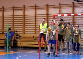 Na zdjęciu widać grupę uczestników stojących przy torze przeszkód w hali sportowej. Na pierwszym planie mężczyzna w zielonej koszulce sportowej i pomarańczowej szarfie przechodzi pomiędzy czerwonymi słupkami, wykonując zadanie konkursowe. Za nim stoją kolejni uczestnicy czekający na swoją kolej. W tle widoczna jest bramka sportowa, drewniane drabinki gimnastyczne oraz ławki.