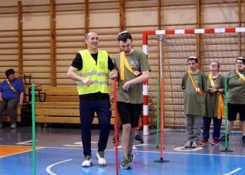 Na zdjęciu widać uczestnika przechodzącego przez tor wyznaczony kolorowymi słupkami w hali sportowej. Towarzyszy mu mężczyzna w żółtej kamizelce odblaskowej. W tle stoją kolejni uczestnicy ubrani w zielone koszulki i pomarańczowe szarfy, czekający na swoją kolej. Za nimi znajduje się bramka sportowa oraz drewniane drabinki gimnastyczne.