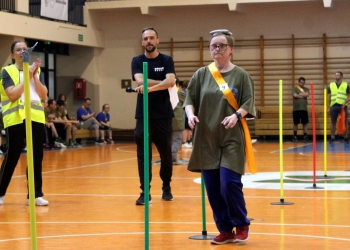 Na zdjęciu widać kobietę uczestniczącą w konkurencji sportowej. Idzie skupiona między kolorowymi słupkami ustawionymi w formie slalomu. Ma na sobie zieloną koszulkę z numerem 9 oraz pomarańczową szarfę przewieszoną przez ramię. Na głowie utrzymuje mały woreczek. W tle znajduje się prowadzący z mikrofonem oraz inni uczestnicy.