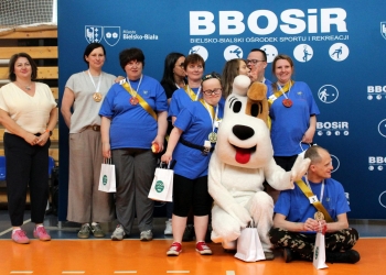 Na zdjęciu widać grupę uczestników wydarzenia sportowego pozujących do wspólnego zdjęcia na hali sportowej. Uczestnicy stoją przed dużą niebieską ścianką z logo BBOSiR oraz napisem „Bielsko-Bialski Ośrodek Sportu i Rekreacji”.  Większość osób ma na sobie niebieskie koszulki, medale zawieszone na szyi oraz kolorowe szarfy z numerami. Niektórzy trzymają w rękach drobne upominki w białych torbach. W centrum zdjęcia znajduje się osoba w dużym kostiumie psa reksia, która pozuje razem z uczestnikami i macha do aparatu.