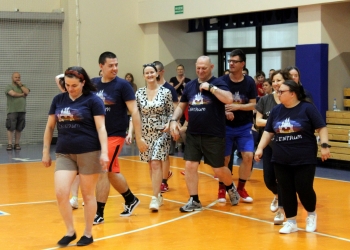 Na zdjęciu widać grupę uczestników idących po hali sportowej. Większość osób ma na sobie granatowe koszulki z napisem „Centrum” i kolorową grafiką na przodzie. Uczestnicy idą razem w luźnym szyku, uśmiechają się i rozmawiają.