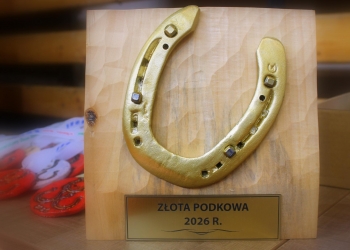 Na zdjęciu znajduje się dekoracyjna złota podkowa przymocowana do jasnej drewnianej tablicy. Podkowa ma metaliczny połysk i ozdobne detale przypominające gwoździe lub kamienie. Na dole tablicy umieszczono złotą plakietkę z napisem „ZŁOTA PODKOWA 2026 R.” W tle, po lewej stronie, widać lekko rozmyte czerwone medale z motywem podkowy oraz białe tasiemki.