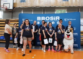 Na zdjęciu widać grupę uczestników wydarzenia sportowego stojących przed dużą niebieską ścianką z logo BBOSiR oraz napisem „Bielsko-Bialski Ośrodek Sportu i Rekreacji”. Większość osób ma na sobie granatowe koszulki z napisem „Centrum”, a na szyjach medale. Niektórzy uczestnicy trzymają białe torby z upominkami. Po prawej stronie klęczy osoba przebrana za psa Reksia.