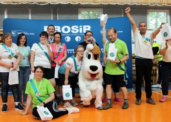 Na zdjęciu widoczna jest grupa uczestników wydarzenia sportowego. Wszyscy stoją i pozują do wspólnego zdjęcia na tle dużej niebieskiej ścianki z napisem „BOSiR” oraz logo miasta Bielsko-Biała. Uczestnicy mają na szyjach medale, a część z nich trzyma dyplomy lub torby z upominkami. Kilka osób unosi ręce w geście radości i świętowania. W centrum zdjęcia znajduje się osoba w dużym kostiumie psa Reksia. 