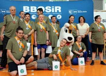 Grupa uczestników wydarzenia sportowego pozujących do wspólnej fotografii w hali sportowej. Wszyscy mają na sobie zielone koszulki, medale na szyjach oraz pomarańczowe szarfy z numerami startowymi. Część osób trzyma białe torby z upominkami.  W centrum grupy znajduje się maskotka dużego psa Reksia. Niektórzy uczestnicy stoją, inni klęczą lub siedzą na podłodze, uśmiechając się do aparatu. W tle widoczna jest duża niebieska ścianka z napisem „BBOSiR” oraz logo miasta Bielsko-Biała.