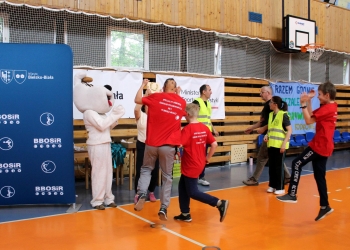 Na zdjęciu uchwycono radosny moment podczas wydarzenia sportowego. Kilka osób w czerwonych koszulkach biegnie i klaszcze z entuzjazmem w kierunku stanowiska z nagrodami lub dekoracją. Jedna z osób unosi ręce w geście zwycięstwa i radości. Po lewej stronie stoi maskotka psa Reksia. Obok znajdują się organizatorzy w żółtych kamizelkach odblaskowych. W tle widoczne są banery z logo „BBOSiR”, miasta Bielsko-Biała oraz inne transparenty związane z imprezą.