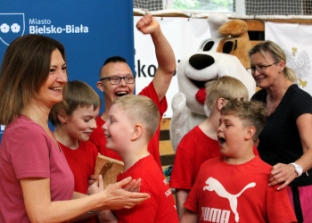 Na zdjęciu widać grupę dzieci i opiekunów podczas radosnego momentu w hali sportowej. Większość uczestników ma na sobie czerwone koszulki. W centrum znajduje się chłopiec, który z entuzjazmem podnosi rękę w geście zwycięstwa i szeroko się uśmiecha. Obok stoją inne dzieci, również wyraźnie podekscytowane i radosne. Po lewej stronie kobieta w różowej bluzce klaszcze, a po prawej druga opiekunka uśmiecha się. W tle widoczna jest duża maskotka psa Reksia.