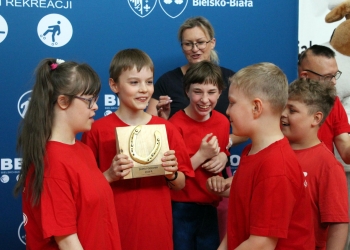 Na zdjęciu widoczna jest grupa dzieci ubranych w czerwone koszulki.W centrum stoi chłopiec trzymający drewnianą nagrodę z symbolem złotej podkowy i napisem „Złota Podkowa 2026 r.”. Dzieci zgromadzone wokół niego uśmiechają się i z zainteresowaniem oglądają trofeum.W tle znajduje się kobieta, która obserwuje grupę z uśmiechem, a za uczestnikami widoczna jest niebieska ścianka z logo „BBOSiR” oraz miasta Bielsko-Biała.  