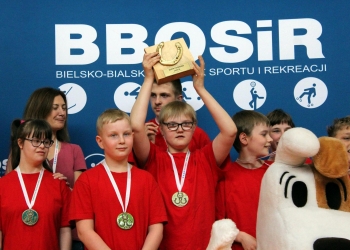 Na zdjęciu widać grupę dzieci i młodzieży ubranych w czerwone koszulki. W centrum stoi chłopiec z medalem na szyi, który unosi nad głową drewniane trofeum z podkową, symbolizujące zwycięstwo. Obok niego stoją inni uczestnicy z medalami. Po lewej stronie znajduje się kobieta opiekująca się grupą, a po prawej widoczna jest duża maskotka psa Reksia. W tle znajduje się niebieska ścianka z logo BBOSiR oraz napisami związanymi z Bielskiem-Białą.