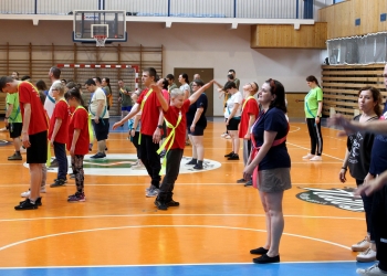 Na zdjęciu widać grupę osób uczestniczących w rozgrywkach sportowych, uczestnicy stoją w rzędach na pomarańczowym boisku i wykonują ćwiczenia ruchowe — część z nich unosi ręce lub rozciąga ciało. Wiele osób ma na sobie kolorowe koszulki, głównie czerwone i zielone, a także przewiązane przez ramię jaskrawe szarfy. W tle widoczne są elementy wyposażenia hali sportowej: kosz do koszykówki, bramki oraz drewniane drabinki gimnastyczne.