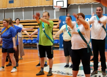Na zdjęciu przedstawiono grupę uczestników biorących udział w ćwiczeniach ruchowych na hali sportowej. Uczestnicy mają na sobie sportowe ubrania w różnych kolorach, m.in. zielonym, białym i niebieskim, a przez ramię przewieszone są kolorowe szarfy z przypiętymi numerami. W tle widoczne są elementy hali gimnastycznej: kosz do koszykówki, siatki zabezpieczające oraz rzędy niebieskich siedzeń.