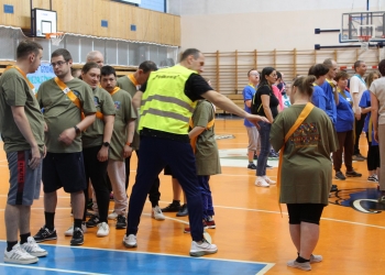 Na zdjęciu widać grupę uczestników zgromadzonych na hali sportowej. Większość osób ubrana jest w oliwkowozielone lub niebieskie koszulki oraz kolorowe szarfy z numerami. W centrum kadru znajduje się mężczyzna w jaskrawej kamizelce odblaskowej z napisem „Podkowa”. W tle widoczne są elementy hali gimnastycznej — kosze do koszykówki, drewniane drabinki oraz siatki zabezpieczające.