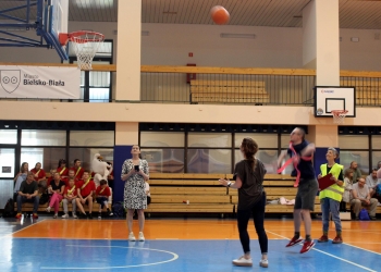 Na zdjęciu uchwycono moment konkurencji sportowej odbywającej się na hali gimnastycznej. W centrum kadru widać lecącą piłkę do koszykówki oraz uczestnika wykonującego rzut w kierunku kosza. Wokół boiska stoją inni uczestnicy i organizatorzy wydarzenia. Jedna z osób ma na sobie żółtą kamizelkę odblaskową i trzyma clipboard, co sugeruje rolę sędziego lub koordynatora. Na trybunach i ławkach w tle siedzą widzowie oraz uczestnicy ubrani w kolorowe koszulki i szarfy.  Na ścianie hali widoczny jest baner z napisem „Miasto Bielsko-Biała”.